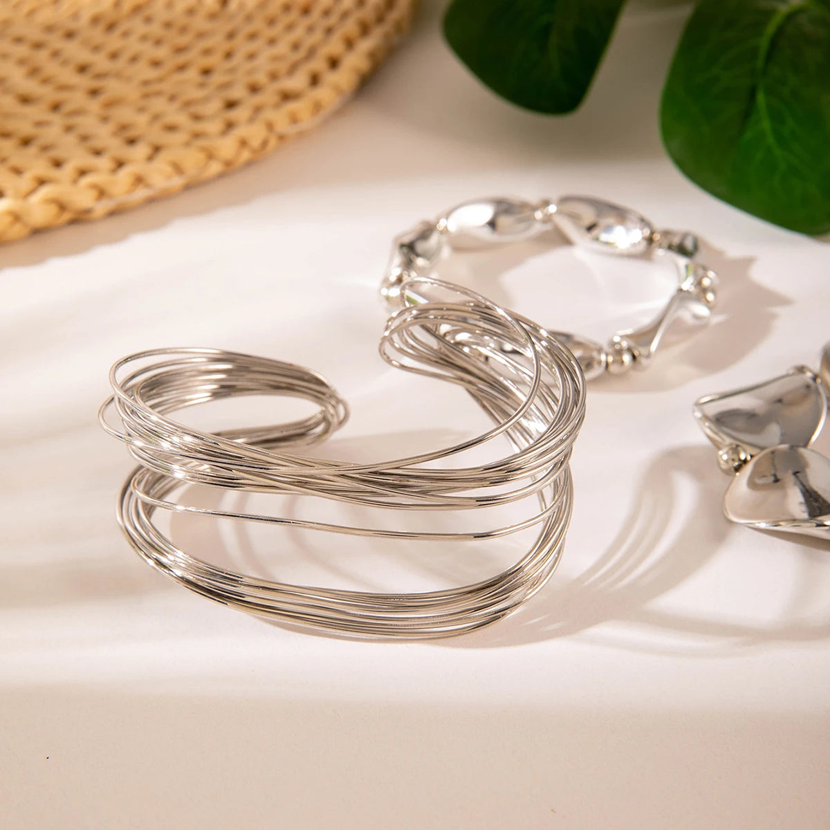 Silver bracelets on a light surface with a woven mat and green leaves in the background