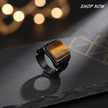 Black ring with a brown gemstone on a dark surface with blurred lights in the background