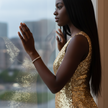 A woman in a gold sequin dress with a diamond bracelet, looking out a window with a blurred cityscape background