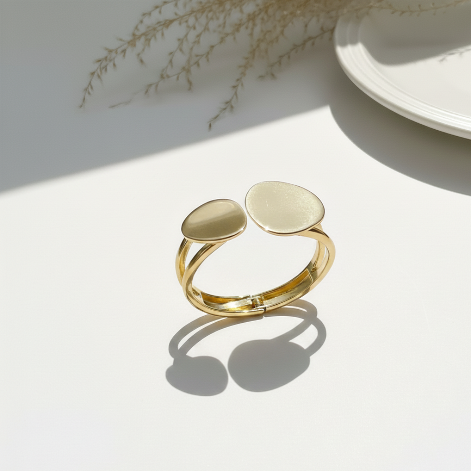 Gold bangle with two oval links pendants on a light surface with a plant in the background