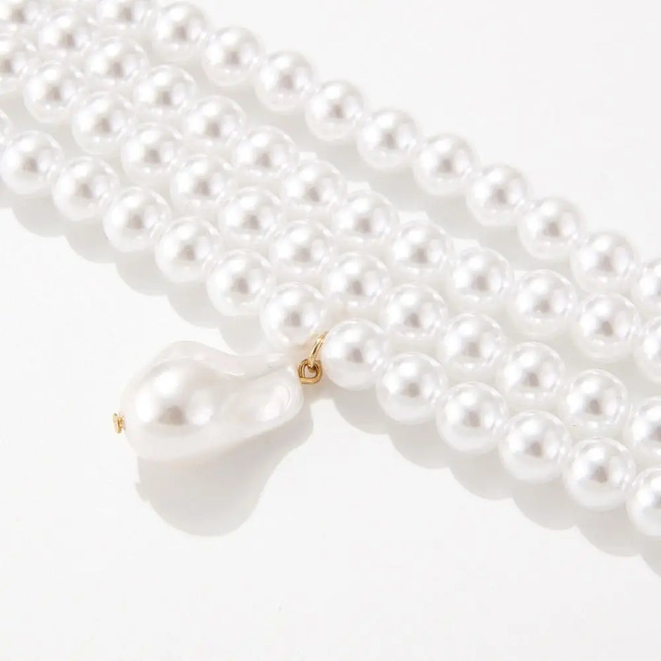 a choker-style necklace made of four rows of small, round white pearls. It has a gold-toned clasp at the back and a single, larger, teardrop-shaped white pearl pendant hanging from the center front.