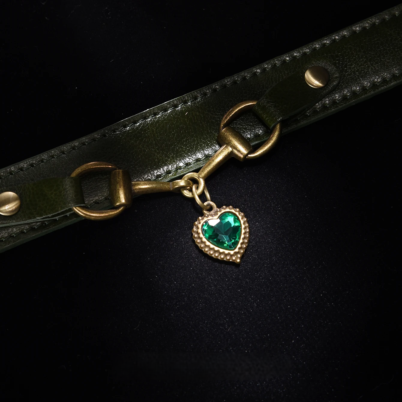 Green leather strap with a gold clasp and heart-shaped emerald green gemstone charm on a black background.