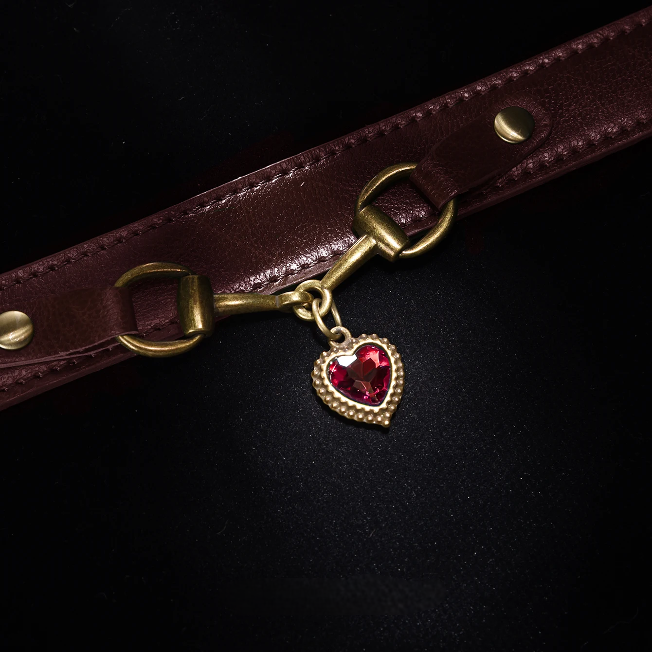 Brown leather strap with a gold buckle and red heart charm on a black background