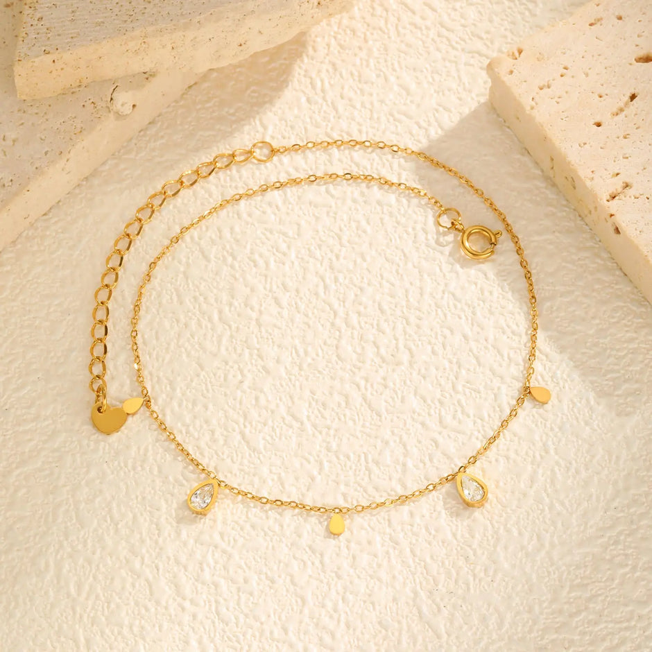 Gold anklet with heart and round charm on a beige surface