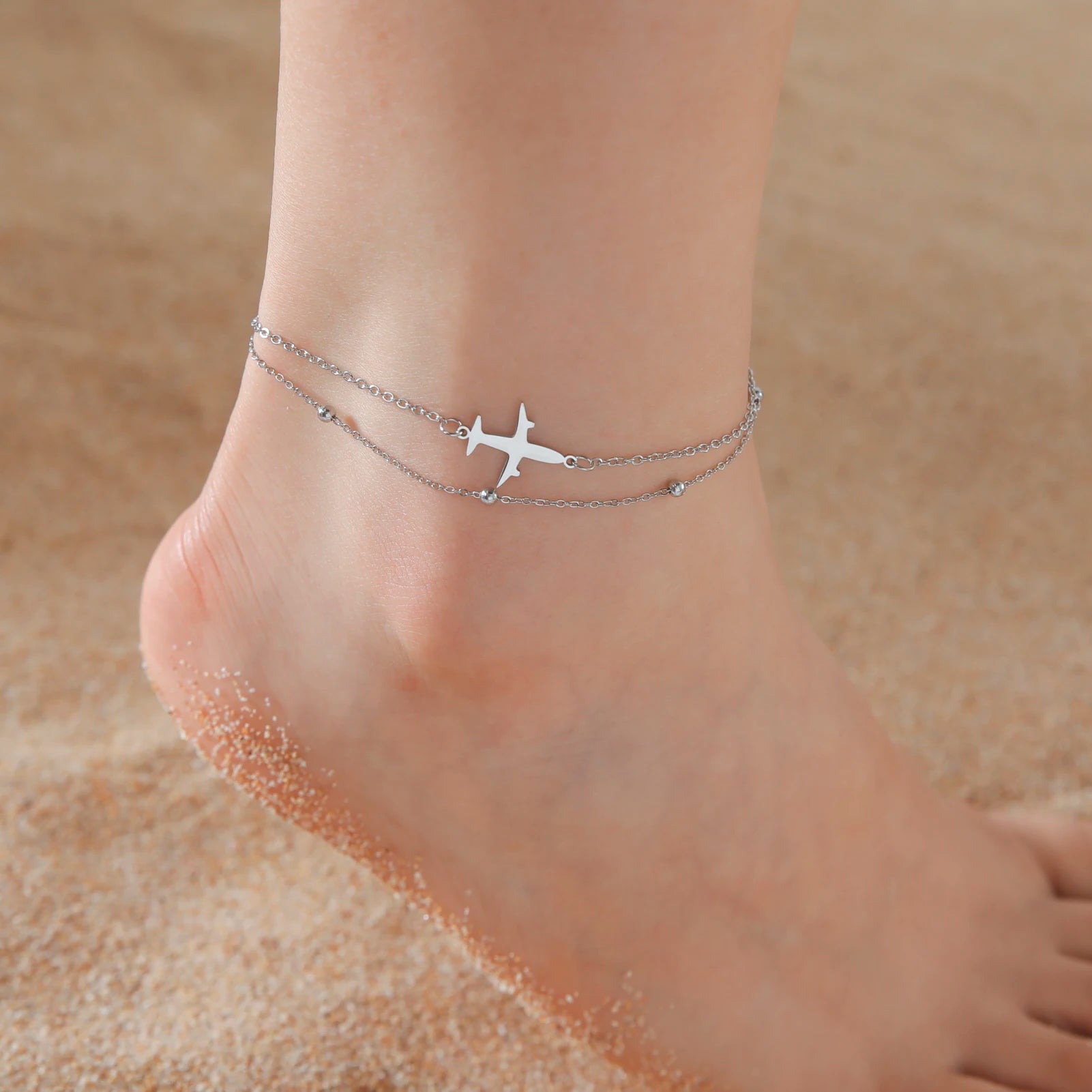 Silver anklet with airplane charm on a foot against a sandy background