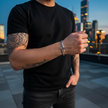Person wearing a black t-shirt with tattoos on arms and hands, standing on a rooftop with city skyline in the background.