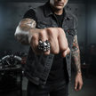 Man wearing a denim vest and skull ring in a dark room with tattoos on arms