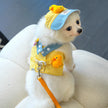 Small white dog wearing a yellow checkered outfit with duck decorations and a blue cap.